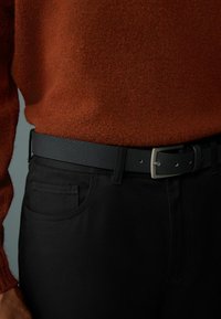 Schwarzer, strukturierter Gürtel mit einer silbernen Schnalle, der über dunklen Hosen getragen wird, kombiniert mit einem figurbetonten, warmen orangefarbenen Pullover.