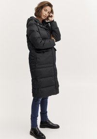 Musthave musta pikk puffer-jakk, millel on kapuuts, tikandiga disain, küljetaskud ja tõmblukk. Kombineeritud siniste džinsidega ja mustade tossudega.