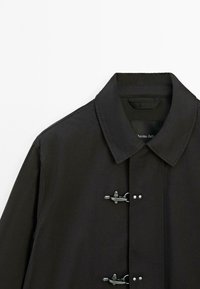 Black coat with a pointed collar, featuring two metal closures and a smooth, flat texture. Label visible at the neck.