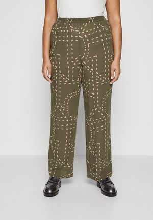 Person iført olivengrønne bukser med beige stiplede abstrakte mønster, hvid top og sorte skinnende loafers, stående mod en ensfarvet baggrund.