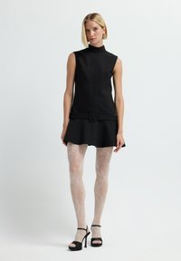 Schwarzes ärmelloses Kleid mit hohem Kragen und Gürteldetail, ergänzt durch transparente weiße gemusterte Strumpfhosen und schwarze High Heels mit offener Zehenpartie.