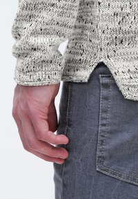 Stickad tröja med en ljus krämfärgad bas, texturerat mönster i svart och beige. Har ribbade ärmslut, rundad nederkant och bärs med grå denim.