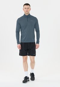 Långärmad, dragkedja sporttop i mörkteal, ihopparad med svarta shorts. Lättviktig, stretchig tyg, enkel design och inga synliga logotyper.