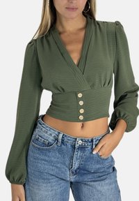 Olivgrüne Bluse mit Wickeldesign, langen Puffärmeln und vier beigen Knöpfen an der Taille. Kombiniert mit hoch taillierter blauer Jeans.