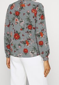 Blumenbluse mit grauem Hintergrund, roten und rosa Blumen, grünen Blättern und langen Ärmeln mit elastischen Bündchen. Taillierter Schnitt mit rundem Ausschnitt.