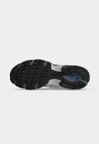 Μαύρη και γκρι σόλα παπουτσιού New Balance με υφή στο πέλμα και δύο μικρές μπλε λεπτομέρειες κοντά στην περιοχή της φτέρνας.