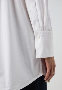 Camicia bianca a maniche lunghe con polsini abbottonati, caratterizzata da una texture liscia e una vestibilità morbida. Cuciture visibili lungo le cuciture.
