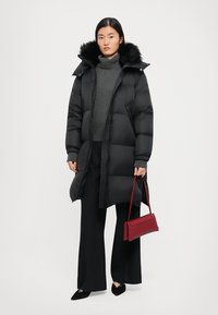 Schwarze Pufferjacke mit einer mit Fell besetzten Kapuze, Reißverschluss und Seitentaschen, getragen über einem dunkelgrauen Pullover und kombiniert mit weiten Hosen und einer roten Handtasche.