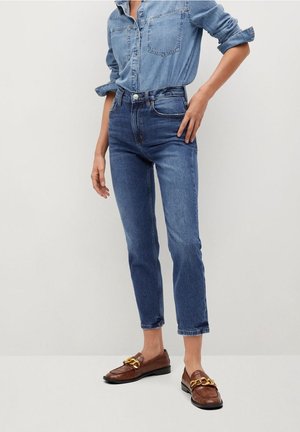 Personne portant une chemise en jean bleue et un jean avec les manches retroussées et des mocassins marron avec un détail chaîne dorée, debout contre un mur clair.