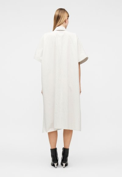 Frau mit langen blonden Haaren, die ein locker sitzendes, knielanges weißes Kleid mit kurzen Ärmeln und schwarze Knöchelstiefel trägt und mit dem Rücken zum Betrachter steht.