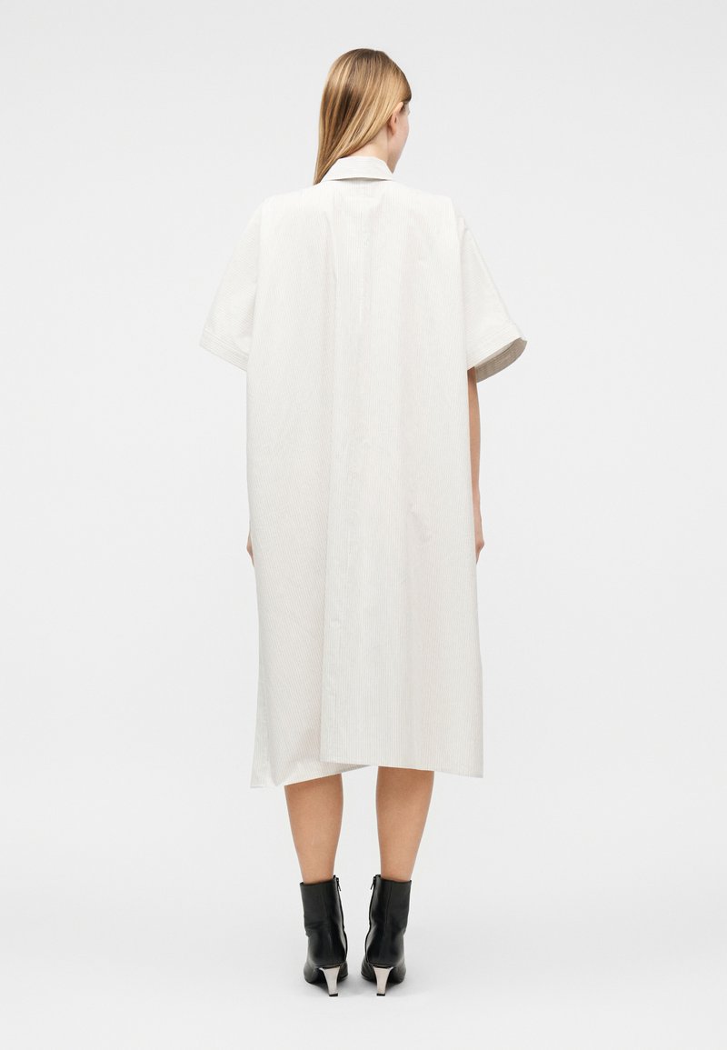 Femme aux longs cheveux blonds portant une robe blanche ample jusqu'aux genoux avec des manches courtes et des bottines noires, debout de dos.