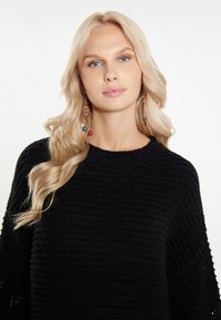 Černý pletený svetr s ribbovaným vzorem, nošený s barevnými korálkovými náušnicemi se zlatými prvky. Dlouhé, vlnité blond vlasy.