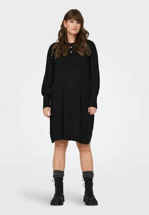 Robe-sweater noire en tricot avec des manches longues et des poignets côtelés, présentant une coupe ample. Associée à des chaussettes grises en tricot et des bottes noires à lacets.