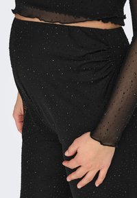 Mustast kangast rõivastus läbipaistva, täpilise tekstuuriga, millel on väiksed helmed; kehakuju järgiv disain, vöökoha kokkutõmmetega ja laineliste varrukaäärtega.