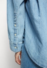 Ljusblå denimskjorta med rundad nederkant, knäppta ärmslut och strukturerad tyg. Ärmen har fem knappar med spetsig kant.