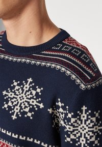 Maglione lavorato a maglia blu navy con motivi di fiocchi di neve bianchi e rossi, collo a costine e una finitura texturizzata intorno alle spalle.