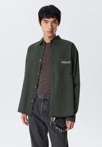 Young man wearing dark green button-up shirt over brown and black striped tee, dark jeans with keychain accessory, standing against white background.