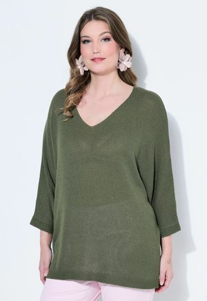 Vrouw met een losse olijfgroene V-hals trui, roze opvallende oorbellen en lichtroze broek tegen een effen witte achtergrond.