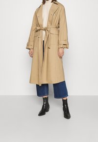 Trench coat beige chiaro con vita a cintura, bottoni e tasche laterali, abbinato a jeans blu a vita bassa e stivaletti alla caviglia. Tessuto dalla trama liscia.