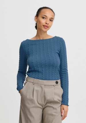 Blau strukturiertes, langärmliges Oberteil mit weiter Halsöffnung, kombiniert mit beigen, gestreiften, hochgeschnittenen Hosen mit schwarzen Knopfdetails.