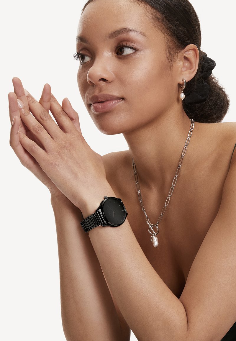 Zwart metalen horloge met een ronde wijzerplaat en een schakelband, gedragen om een pols, naast een zilveren ketting met een parel als hanger.
