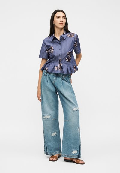 Femme portant un chemisier bleu floral à manches courtes avec un bas peplum, un jean large déchiré et des sandales marron à bout ouvert, debout sur un fond blanc.