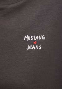 Tmavosivá látka s bielym vyšívaným textom "MUSTANG" nad červeným srdcom a "JEANS" pod srdcom, umiestnené v strede materiálu.