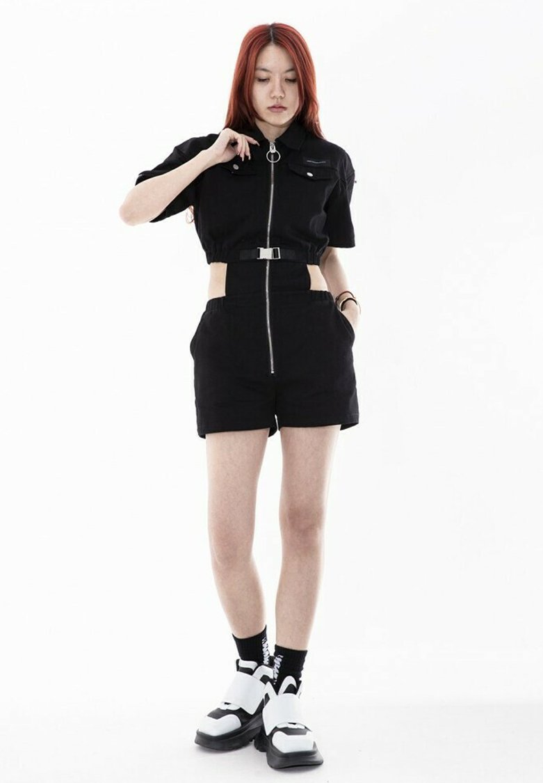 Camiseta negra corta con cremallera en la parte delantera y detalle de anillo, combinada con pantalones cortos negros de talle alto y zapatos gruesos blanco y negro.