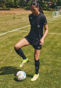 Svart treningsskjorte og shorts med logo, svarte sokker og livlige grønne fotballsko. Fotball med fargerike design på gressfelt.