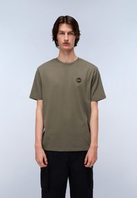 Tricou din bumbac de culoare verde măslin, cu guler rotund, mâneci scurte și un mic logo circular pe pieptul stâng. Combinat cu pantaloni negri.