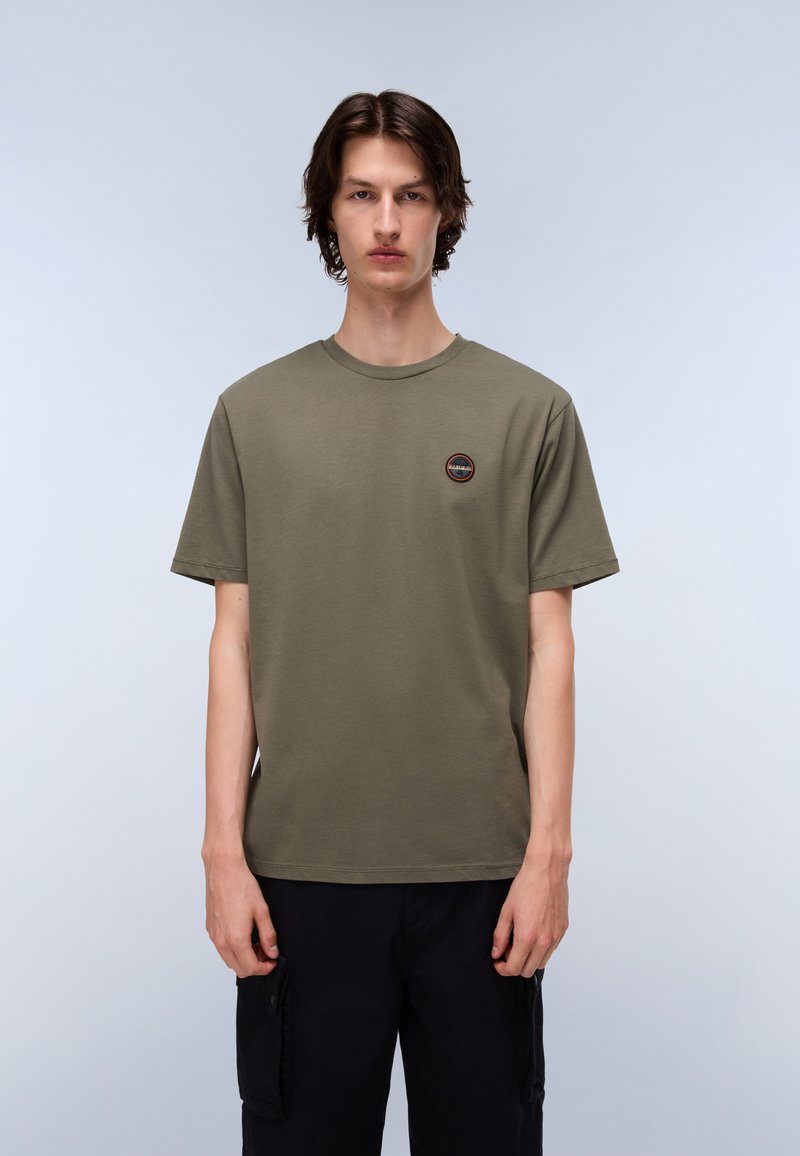 Tricou din bumbac de culoare verde măslin, cu guler rotund, mâneci scurte și un mic logo circular pe pieptul stâng. Combinat cu pantaloni negri.