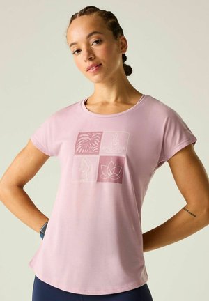 Young woman wearing a pink t-shirt with yoga and nature icons, standing with hands on hips, against a plain light background.