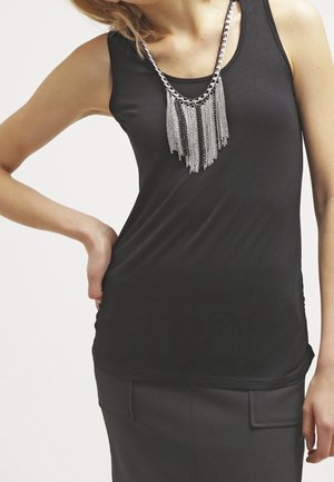 Femme portant un haut noir sans manches, une jupe noire et un collier chaîne argentée avec des détails de franges suspendues.