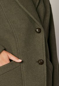Abrigo de lana verde oliva con diseño de doble botonadura, grandes botones texturizados y un cuello ancho. Cuenta con bolsillos laterales y una textura suave.