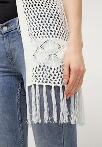 Offenes, gestricktes weißes Poncho mit geometrischen Mustern, ausgestattet mit langen Fransen am Saum, getragen über einem schwarzen Top und blauen Jeans.