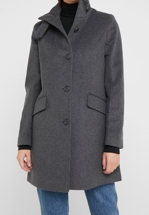 Manteau en laine gris longueur genou avec fermeture à boutons, col cranté, poches à rabat, porté sur un col roulé noir et un jean bleu.