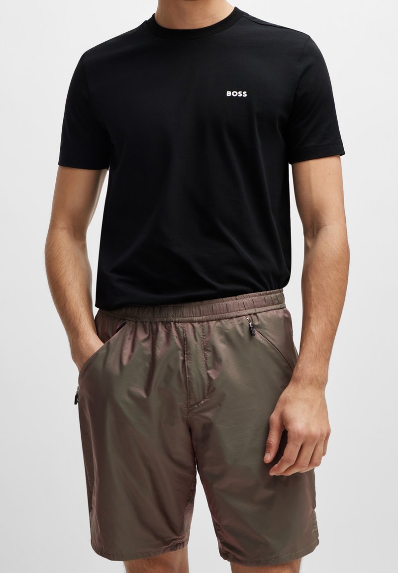 Homme portant un T-shirt noir à manches courtes avec le texte "BOSS" et un short marron à taille élastique avec poches zippées, debout, les mains dans les poches.