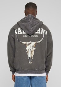 Szürke kapucnis pulóver, nagy bika koponya grafikával a hátsó oldalon, fehér "KAIMAN EST. 1989" felirattal felette. Puha, textúrált anyag.