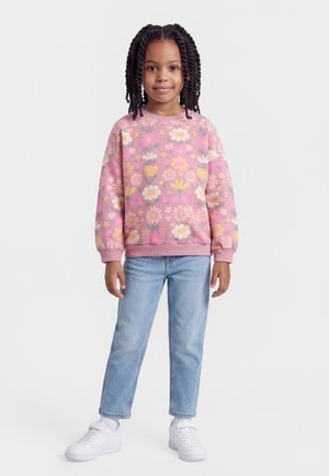 Kind mit lockigem Haar, trägt ein rosa Sweatshirt mit Blumenmuster, hellblaue Jeans und weiße Turnschuhe, steht vor einem schlichten Hintergrund.