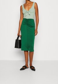 Body côtelé vert menthe avec un détail nœud à l'avant, associé à une jupe midi ajustée vert foncé à fronces sur les côtés, et un sac à main noir.
