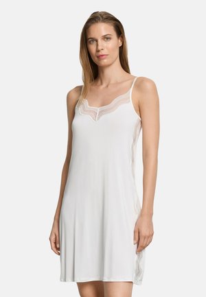 Kvinde iført en hvid ærmeløs slipkjole med blondekant langs halsudskæringen og siderne, stående mod en ensfarvet lys baggrund.