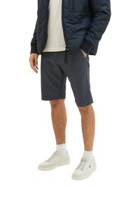 Marineblau gemusterte Shorts mit lässigem Schnitt, aus strukturiertem Stoff, kombiniert mit einem hellen Hemd und weißen Sneakers.