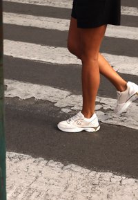 Vita atletiska sneakers i vitt med en strukturerad yta, som har en metallisk guldaccent och en logotyp. Bäraren sätter foten på ett övergångsställe.