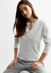 Cardigan grigio chiaro, design con bottoni e scollatura rotonda, polsini a coste e texture morbida. Il modello è seduto, indossando pantaloni grigi.