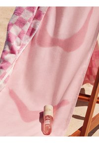 Serviette de plage rose à motif à carreaux sur le sable, chaise longue en bois et une petite bouteille d'huile parfumée Sol de Janeiro Cheeky Bikini.