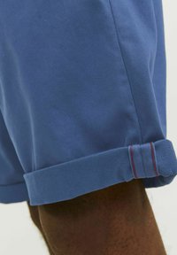 Shorts en coton bleu avec un ourlet roulé, présentant un détail de couture rouge contrasté. Le matériau est lisse, avec une coupe ajustée au niveau de la cuisse.