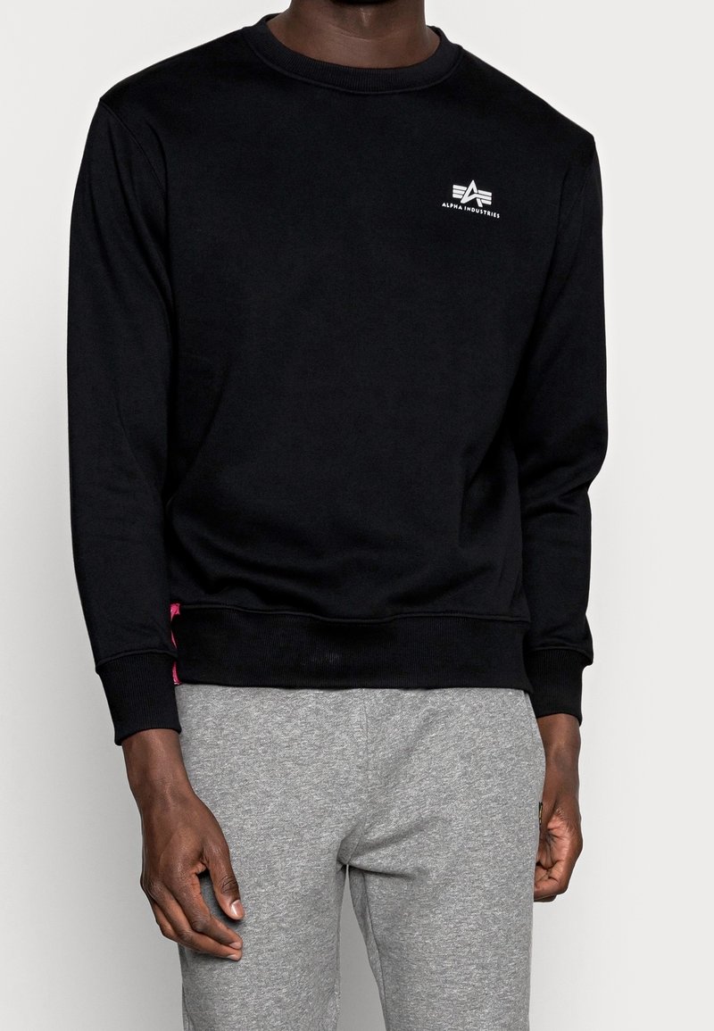 Sweat-shirt noir avec un col rond, arborant un petit logo blanc sur la poitrine et des accents roses à l'ourlet latéral. Tissu doux et texturé.