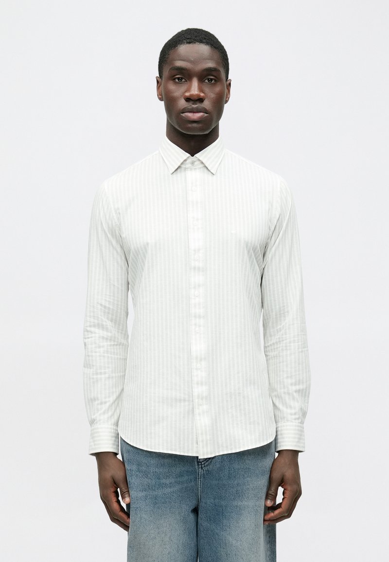 Weißes Langarmhemd mit hellen vertikalen Streifen und einem Button-Down-Kragen, aus glattem Stoff gefertigt. Wird mit hellblauen Jeans getragen.