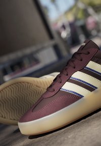 Baskets Adidas bordeaux et crème avec des bandes bleues et une semelle en gomme, placées à l'extérieur sur une surface en béton sous une lumière douce du jour.