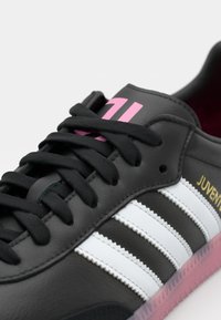 Fekete bőr sneaker fehér csíkokkal, fekete fűzőkkel és rózsaszín akcentussal. Texturált felülettel és áttetsző rózsaszín talppal rendelkezik.
